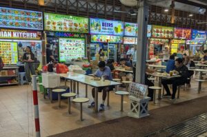 Wide shot of a busy Singaporean hawker center interior featuring various food stalls, including Indian and Thai cuisine, with locals dining at communal tables.
