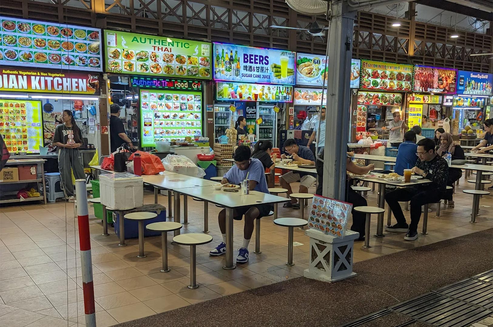 Wide shot of a busy Singaporean hawker center interior featuring various food stalls, including Indian and Thai cuisine, with locals dining at communal tables.