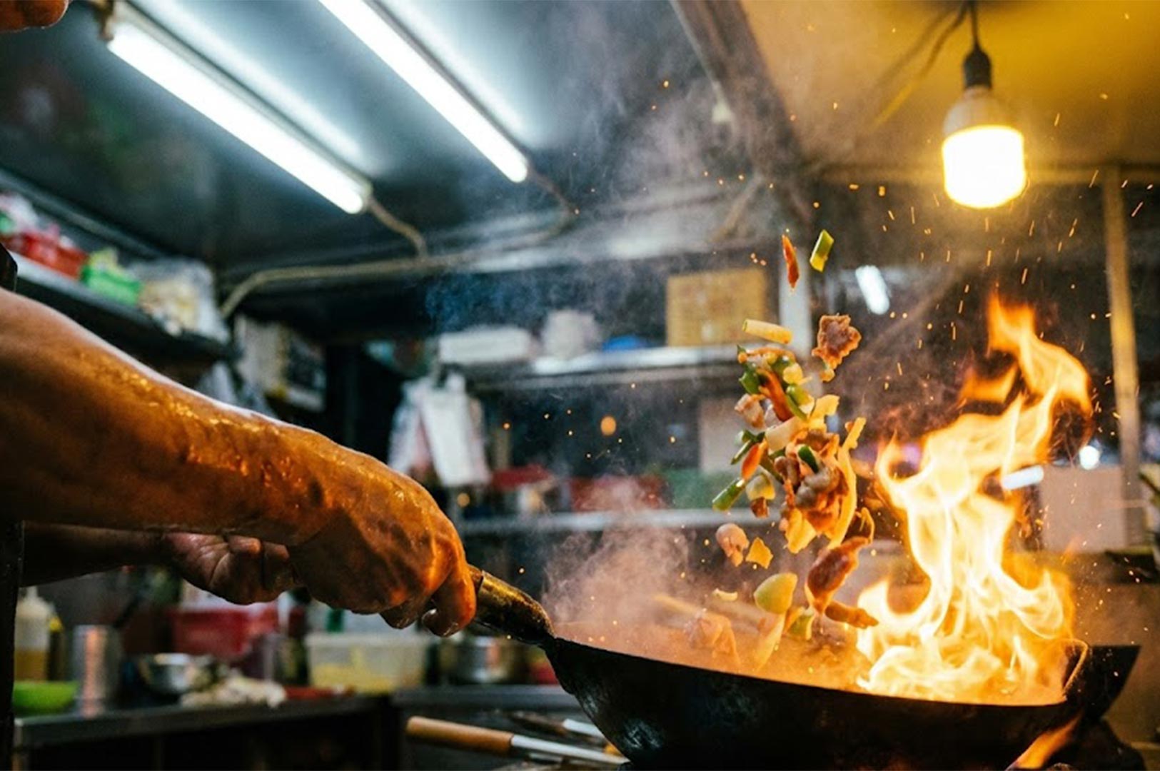 Action shot of a street food chef tossing fresh ingredients in a wok with a large burst of fire, demonstrating the "wok hei" cooking technique.