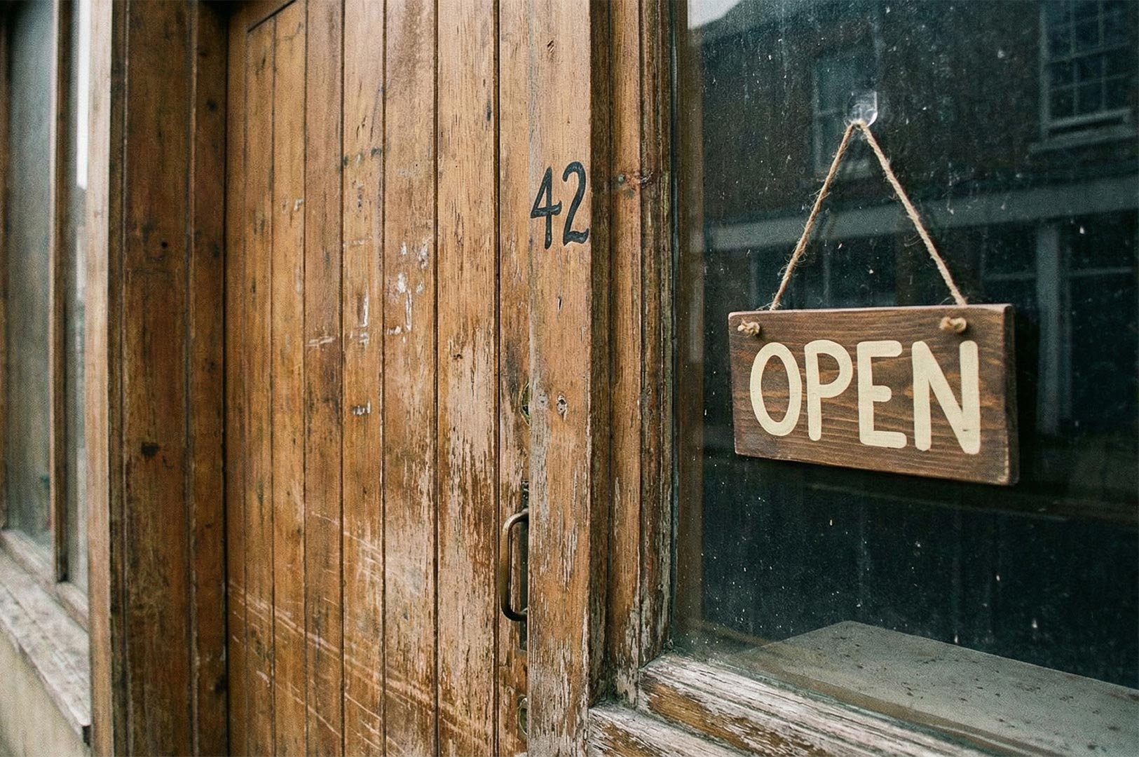 A rustic, rectangular wooden sign reading "OPEN" hanging by string on a weathered wooden door marked with the number 42.