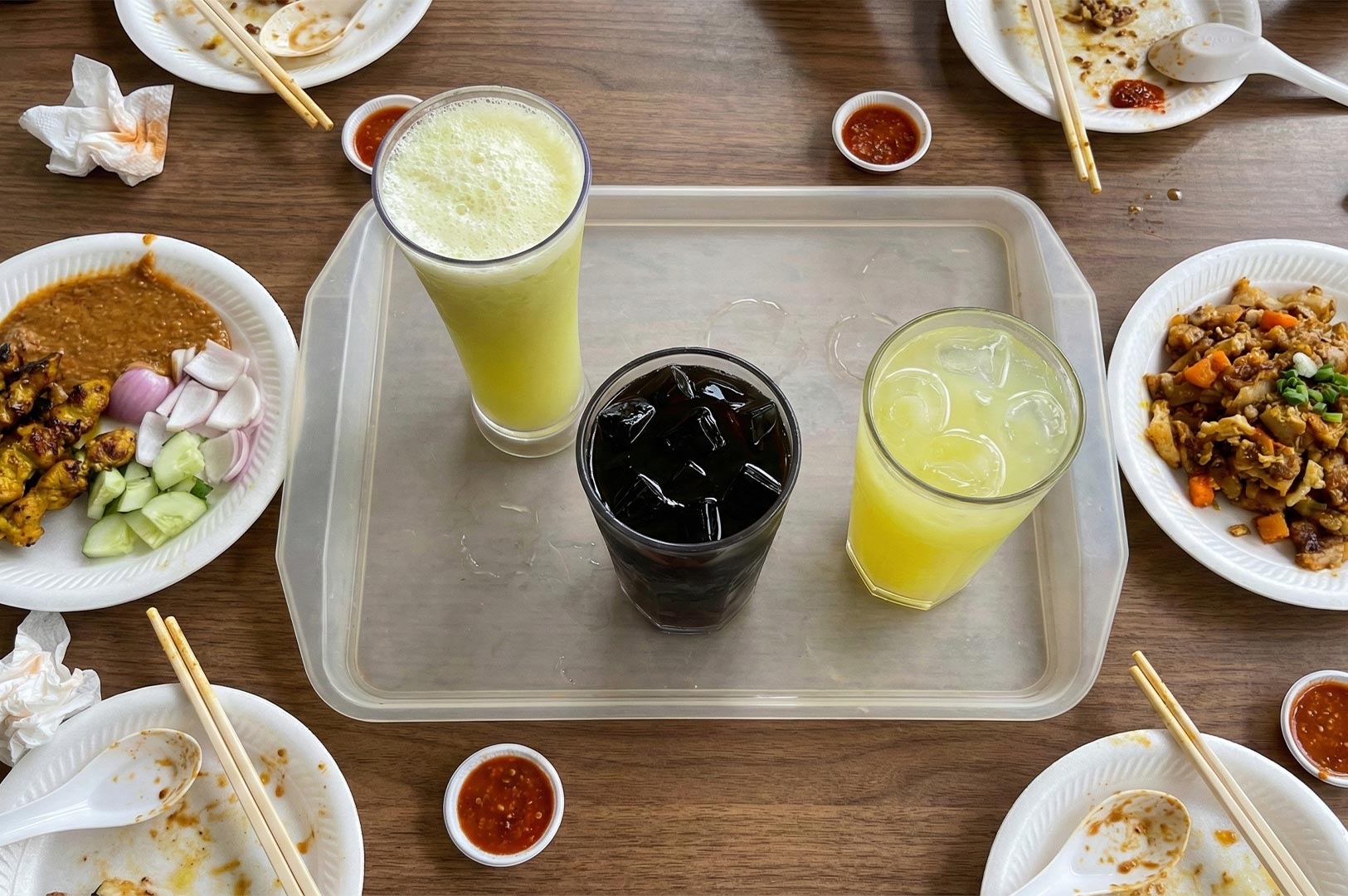 Top-down view of a food tray featuring fresh sugarcane juice and iced coffee (kopi), surrounded by plates of satay with peanut sauce and fried noodles.