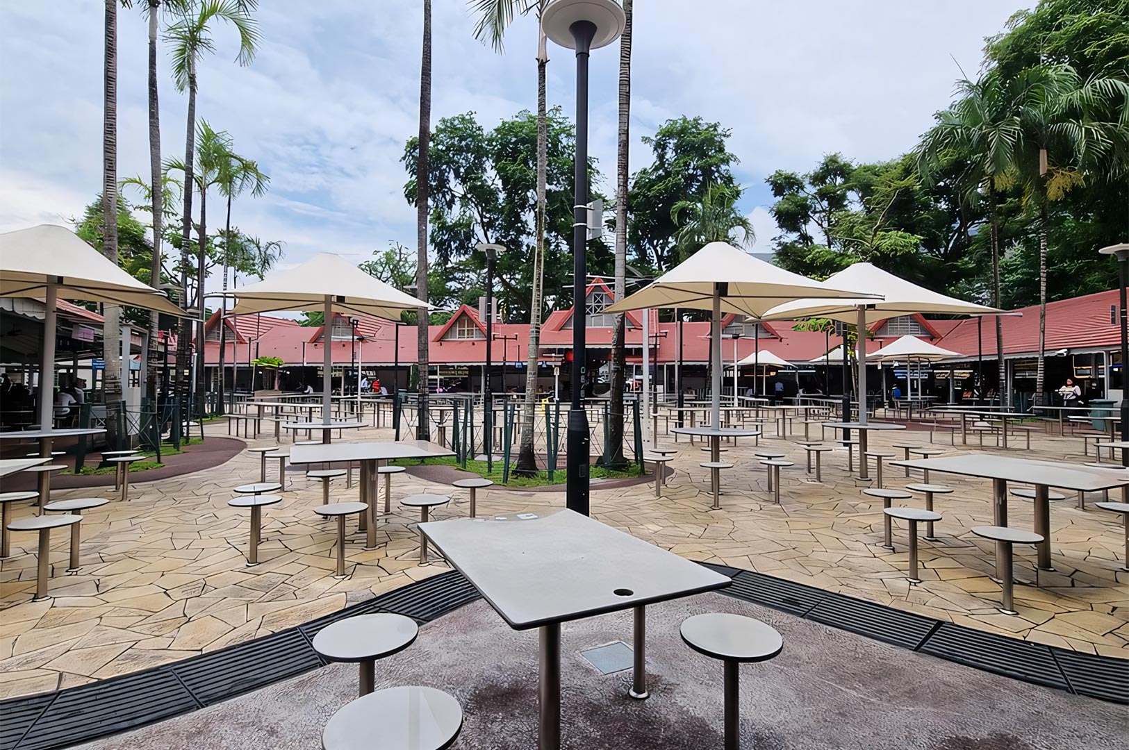 The open-air courtyard of Newton Food Centre during the day, featuring its distinctive colonial-style red roofs, stone tables, and surrounding palm trees.