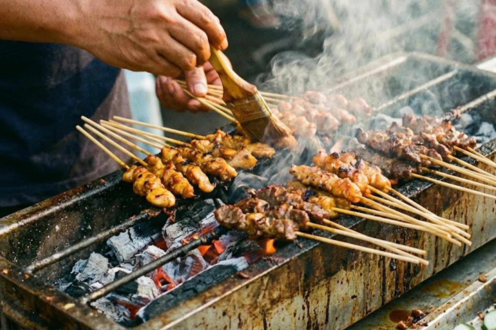 Close-up action shot of satay skewers being brushed with oil and grilled over a smoking open charcoal fire.