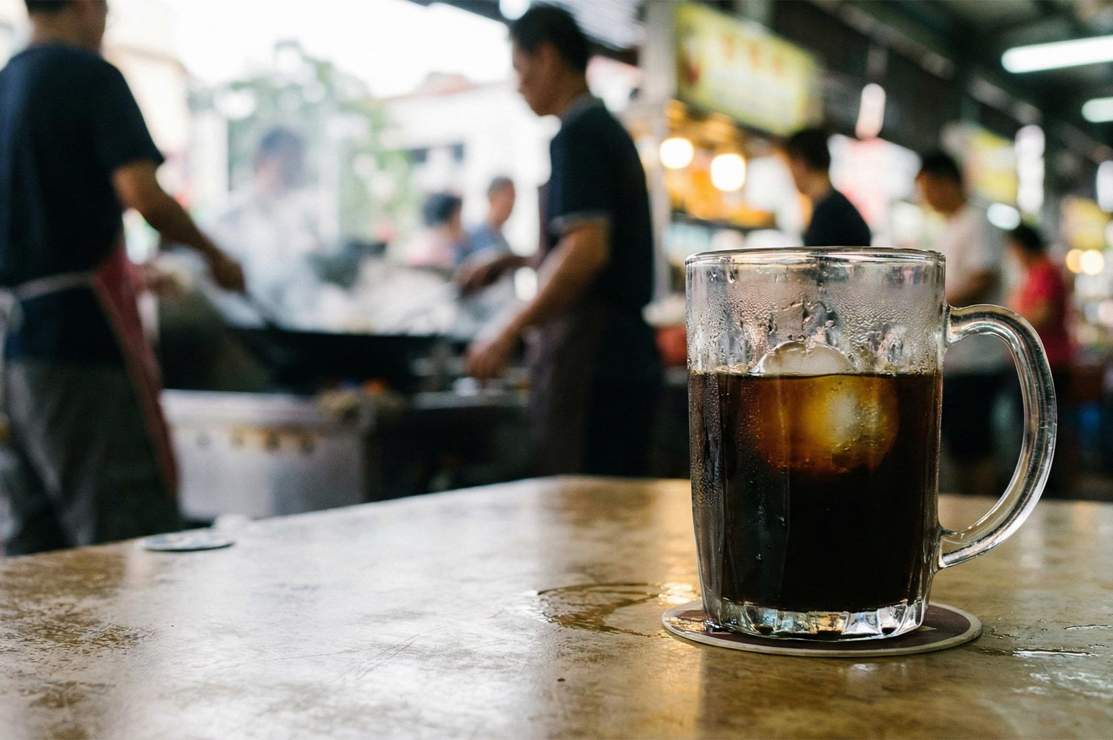 Close-up of a traditional glass mug filled with iced black coffee (Kopi-O), sitting on a table with a blurred background of hawker cooks working in the kitchen.