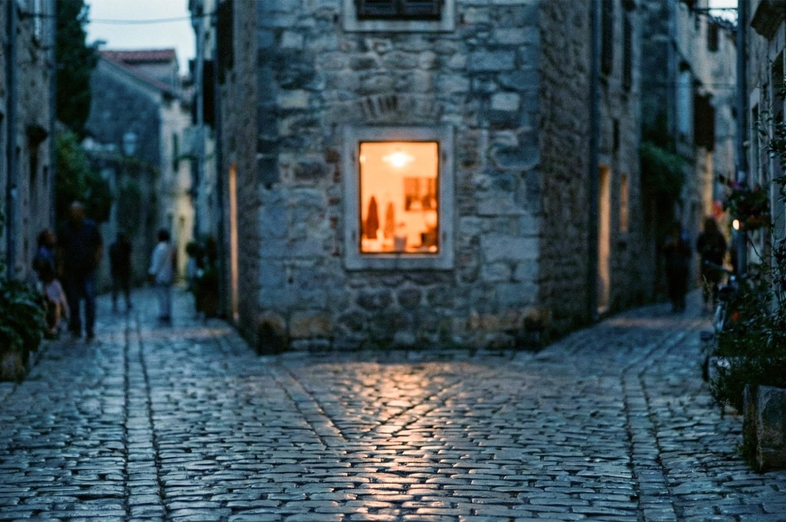 A narrow cobblestone street in a historic European-style town at dusk, featuring old stone buildings and a warm glow emanating from a single square window.