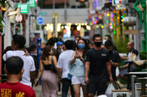 A bustling, narrow outdoor street at night with colorful lights and people walking past open-air bars and restaurants.