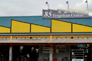 Exterior view of the 85 Fengshan Centre in Singapore, featuring a corrugated blue and yellow facade with its signature signage under a cloudy sky.