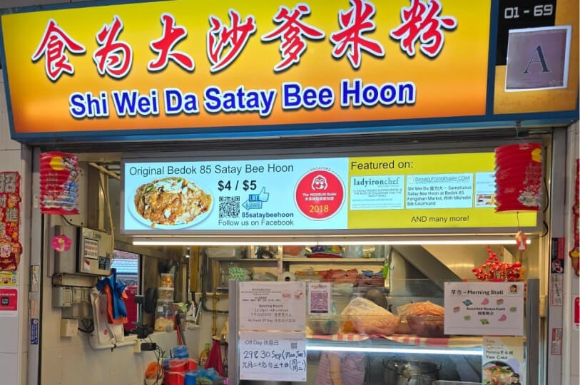 A vibrant yellow storefront of a Singaporean hawker stall named "Shi Wei Da Satay Bee Hoon," featuring Michelin Guide 2018 branding and price signs for its signature noodle dish.