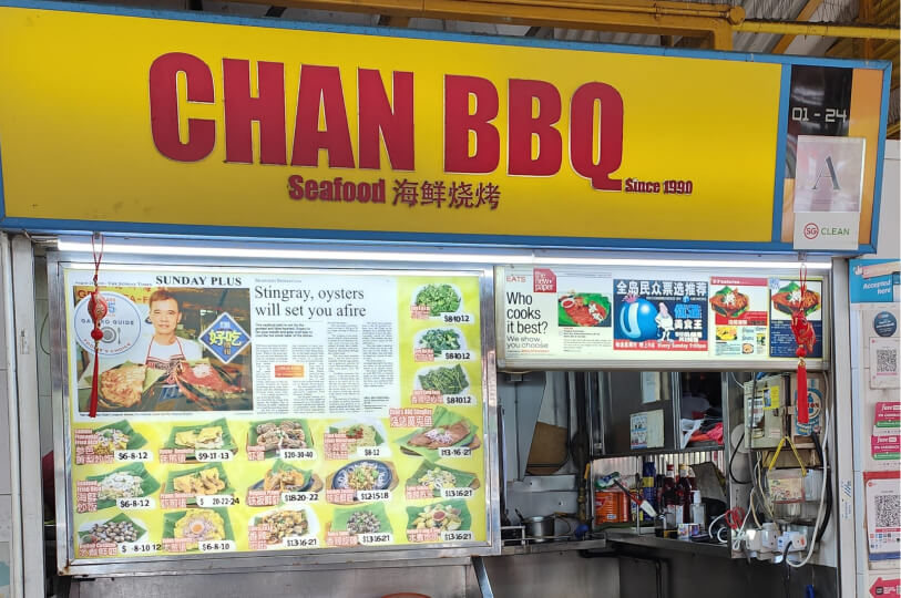 Storefront of the "Chan BBQ Seafood" stall, distinguishable by its bright yellow sign with red lettering and a front display covered in menu photos of seafood dishes and newspaper reviews.