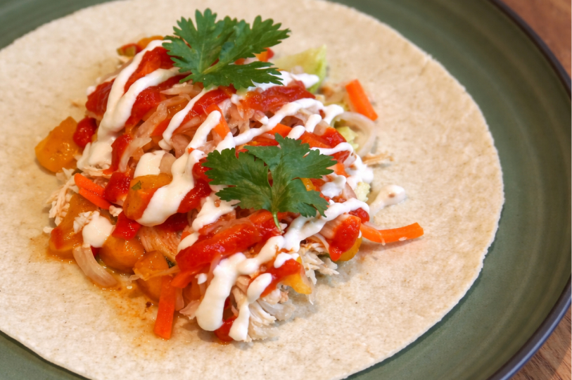 A single open-faced soft taco on a green plate, topped with shredded chicken, peppers, carrots, red salsa, and a drizzle of white cream.