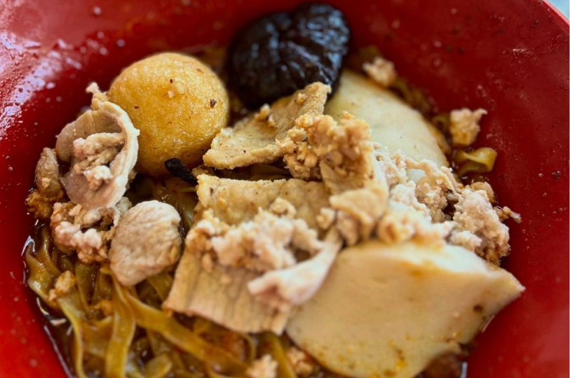 Close-up overhead shot of a bowl of Bak Chor Mee (minced meat noodles), featuring flat yellow noodles topped with minced pork, pork slices, a meatball, and a braised mushroom in a red bowl.