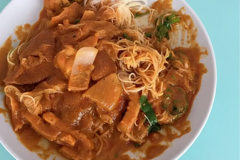 A close-up view of Satay Bee Hoon on a white plate, showing the thin rice noodles heavily coated in a textured, savory peanut gravy with visible pieces of pork and vegetables.