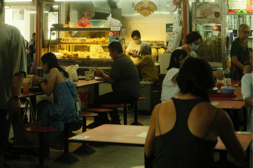 An indoor food court or hawker center with people sitting at tables and a vendor stall in the background displaying golden-fried snacks.