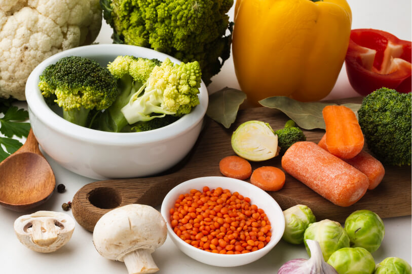 A vibrant assortment of raw ingredients including broccoli, romanesco, bell peppers, carrots, mushrooms, and a bowl of red lentils on a white surface.