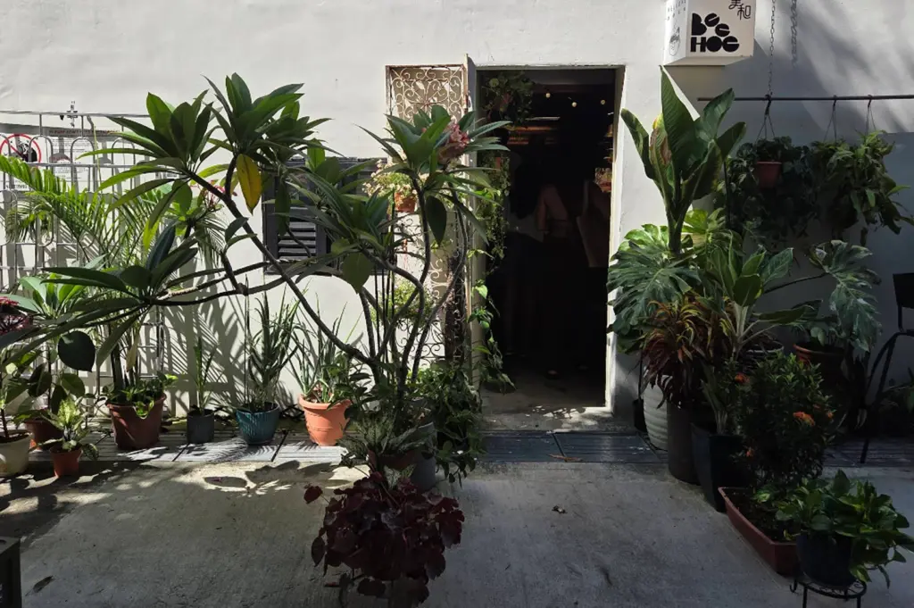 A wide, straight-on shot of the entrance to Bee Hoe Coffee, nestled in a weathered white concrete building. The doorway is framed by an abundant collection of potted tropical plants, including large frangipani trees with lush green leaves and various smaller succulents. To the right of the dark open doorway, a glowing square lightbox sign features the "Bee Hoe" logo and Chinese characters. Harsh, natural sunlight creates deep shadows across the concrete ground, highlighting the organic textures of the foliage and the decorative iron gate to the left.