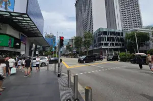 A wide-angle street view of a busy intersection in Singapore's Orchard Road shopping district. Pedestrians wait at a corner under a clear sky. To the left is a modern building with a large digital screen and an Adidas store. Across the street stands a tall, grey skyscraper and a smaller glass-fronted commercial building. Several black and silver cars are moving through the junction, and a red traffic light is visible in the foreground.