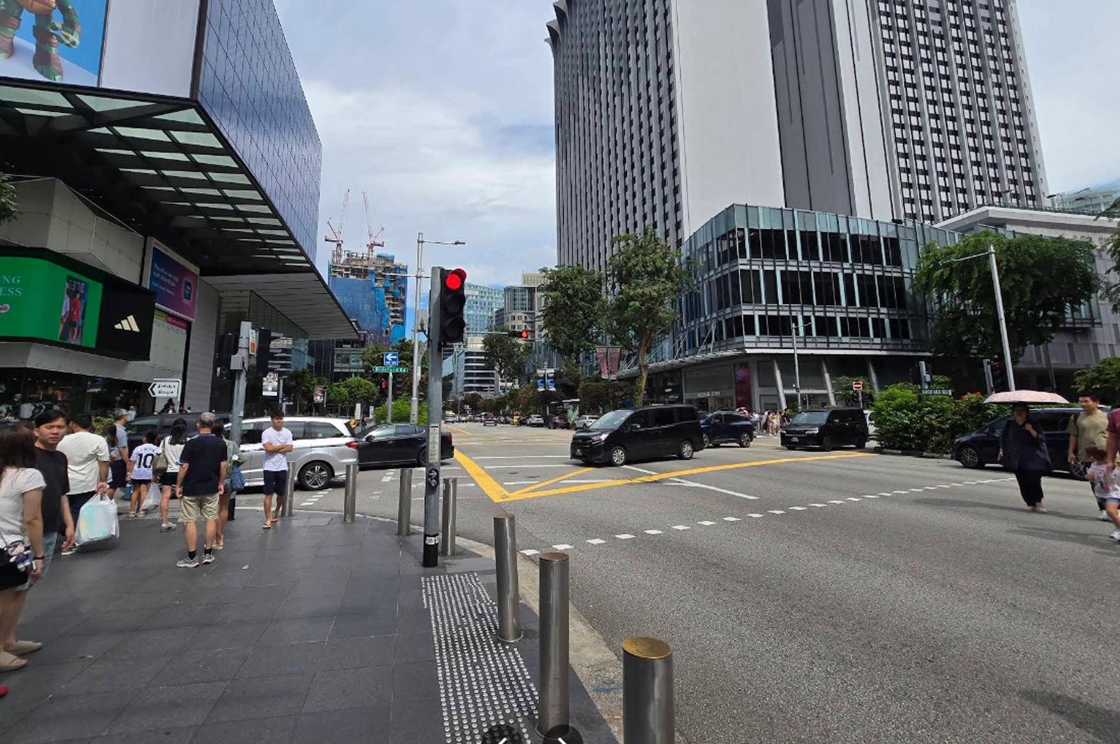 A wide-angle street view of a busy intersection in Singapore's Orchard Road shopping district. Pedestrians wait at a corner under a clear sky. To the left is a modern building with a large digital screen and an Adidas store. Across the street stands a tall, grey skyscraper and a smaller glass-fronted commercial building. Several black and silver cars are moving through the junction, and a red traffic light is visible in the foreground.