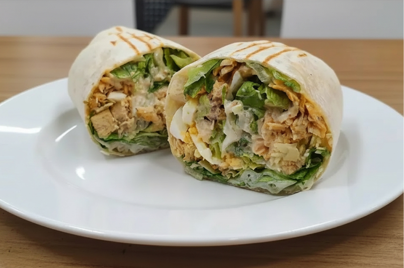A close-up of a grilled tortilla wrap cut in half on a white plate, revealing a dense filling of chicken, boiled eggs, and fresh lettuce.