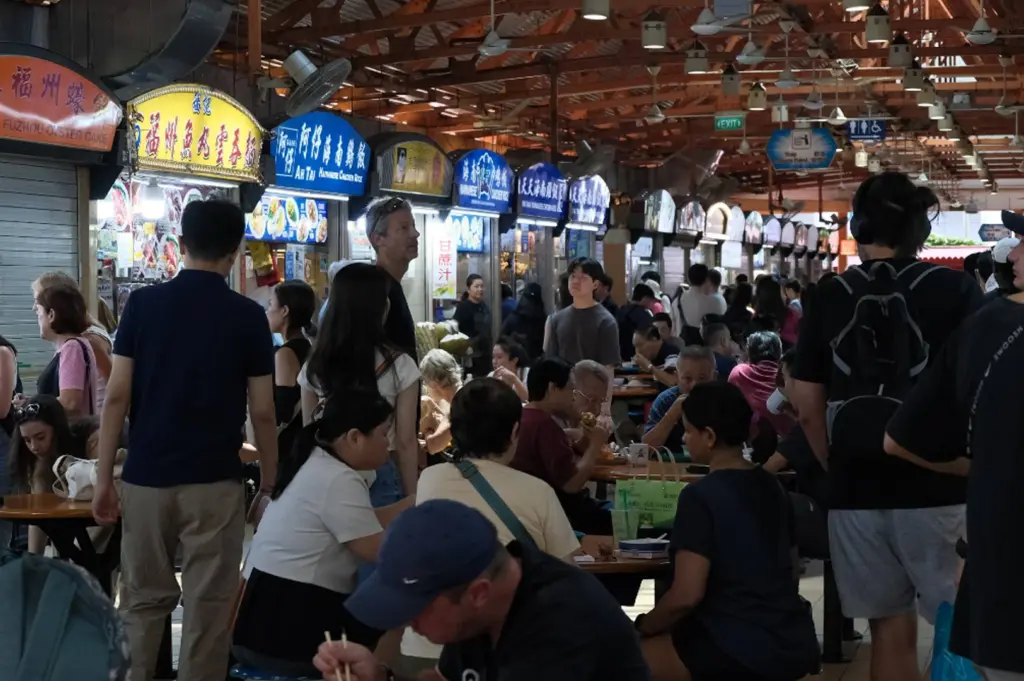 Maxwell Food Centre in Singapore. In the foreground and middle ground, numerous people of diverse backgrounds are seated at circular wooden tables, engaged in conversation while eating. Several people are standing or walking through the narrow aisles between the seating area and a row of hawker stalls. The stalls feature brightly lit, colorful overhead signs in English and Chinese, including "Ah Tai Hainanese Chicken Rice" and "Fuzhou Oyster Cake." The background reveals the industrial-style high ceiling of the food center, equipped with large metal ventilation ducts and multiple rows of white ceiling fans. The lighting is a mix of natural ambient light and the warm glow from the individual food stalls.