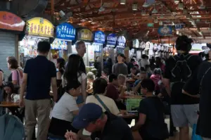 Maxwell Food Centre in Singapore. In the foreground and middle ground, numerous people of diverse backgrounds are seated at circular wooden tables, engaged in conversation while eating. Several people are standing or walking through the narrow aisles between the seating area and a row of hawker stalls. The stalls feature brightly lit, colorful overhead signs in English and Chinese, including "Ah Tai Hainanese Chicken Rice" and "Fuzhou Oyster Cake." The background reveals the industrial-style high ceiling of the food center, equipped with large metal ventilation ducts and multiple rows of white ceiling fans. The lighting is a mix of natural ambient light and the warm glow from the individual food stalls.