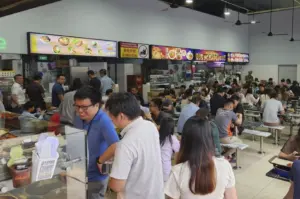 A bustling, crowded indoor food court filled with people dining at long communal tables under bright lights and ceiling fans, with various food stall menus visible in the background.