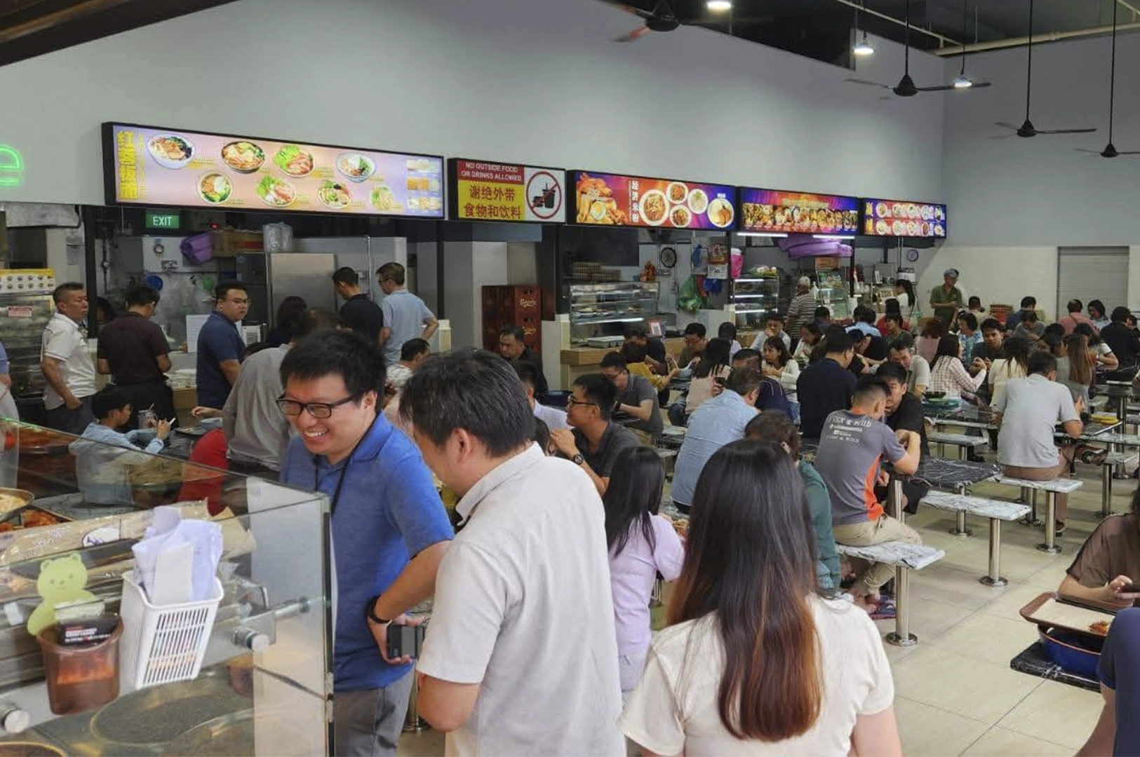 A bustling, crowded indoor food court filled with people dining at long communal tables under bright lights and ceiling fans, with various food stall menus visible in the background.