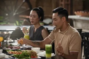 A candid, eye-level shot of a man and a woman enjoying a meal at an outdoor restaurant. The man, in the foreground, wears a tan polo shirt and has a visible tattoo on his forearm as he reaches for a dish with a pair of chopsticks. Beside him, a woman in a grey sleeveless top is engaged in conversation. On the table, there is a spread of various dishes, including fresh greens, noodles, and dipping sauces, alongside two tall glasses of vibrant green juice with metal straws. The background shows a rustic setting with a large wooden wagon wheel and soft, natural lighting.
