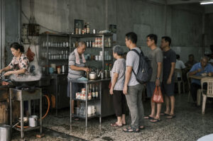 Bustling interior of a rustic, open-air traditional coffee shop or "kopitiam." On the left, a woman in a floral shirt tends to a large, steaming wok on a portable gas stove, while an older man in a striped polo and a white towel around his neck prepares drinks behind a stainless steel counter cluttered with evaporated milk cans, glass jars, and kettles. A patient queue of four people—an elderly woman followed by three younger men in casual attire—stands in the center, waiting to order. The background reveals a dimly lit dining area with weathered concrete walls and patrons seated at round tables, while the speckled grey floor is wet with patches of moisture, adding to the lived-in, authentic atmosphere of the scene.