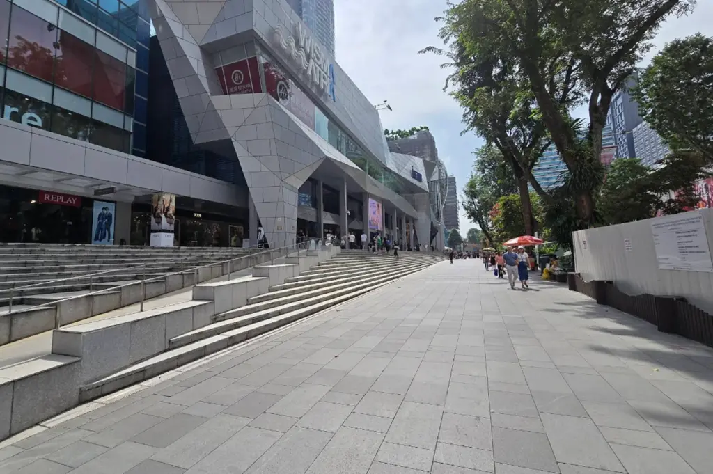 A view of the wide pedestrian walkway outside the Wisma Atria shopping mall. The building features a distinctive, angular grey facade with large glass windows. To the right, tall leafy trees line the promenade, casting shadows on the grey stone tiling. A few people are seen walking in the distance, and a white construction fence is visible on the far right.
