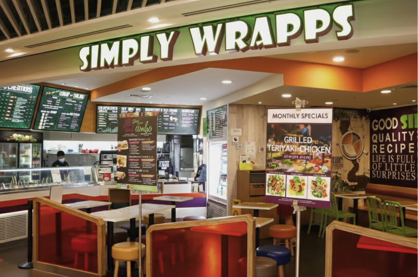 The interior of the Simply Wrapps outlet, showing colorful seating, menu boards, and a "Monthly Specials" poster for Grilled Teriyaki Chicken.