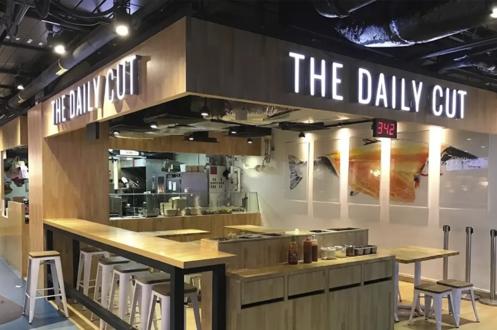 A modern, clean-lined health food eatery called "The Daily Cut" featuring a light wood exterior, minimalist white stools at a high counter, and digital order numbers displayed above the kitchen.