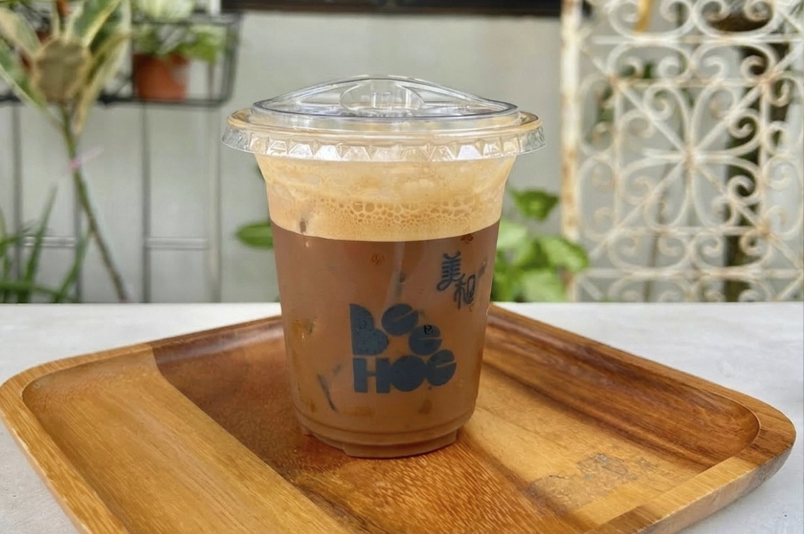 A close-up, eye-level shot of an iced coffee served in a clear plastic cup with a dome lid, sitting on a textured wooden serving tray. The beverage has a distinct gradient, with dark coffee at the bottom rising into a thick, frothy tan foam at the top. The "Bee Hoe" logo is printed in black on the front of the cup. The background is softly blurred, showing hints of garden greenery and a white trellis, keeping the focus on the condensation and textures of the drink and wood.
