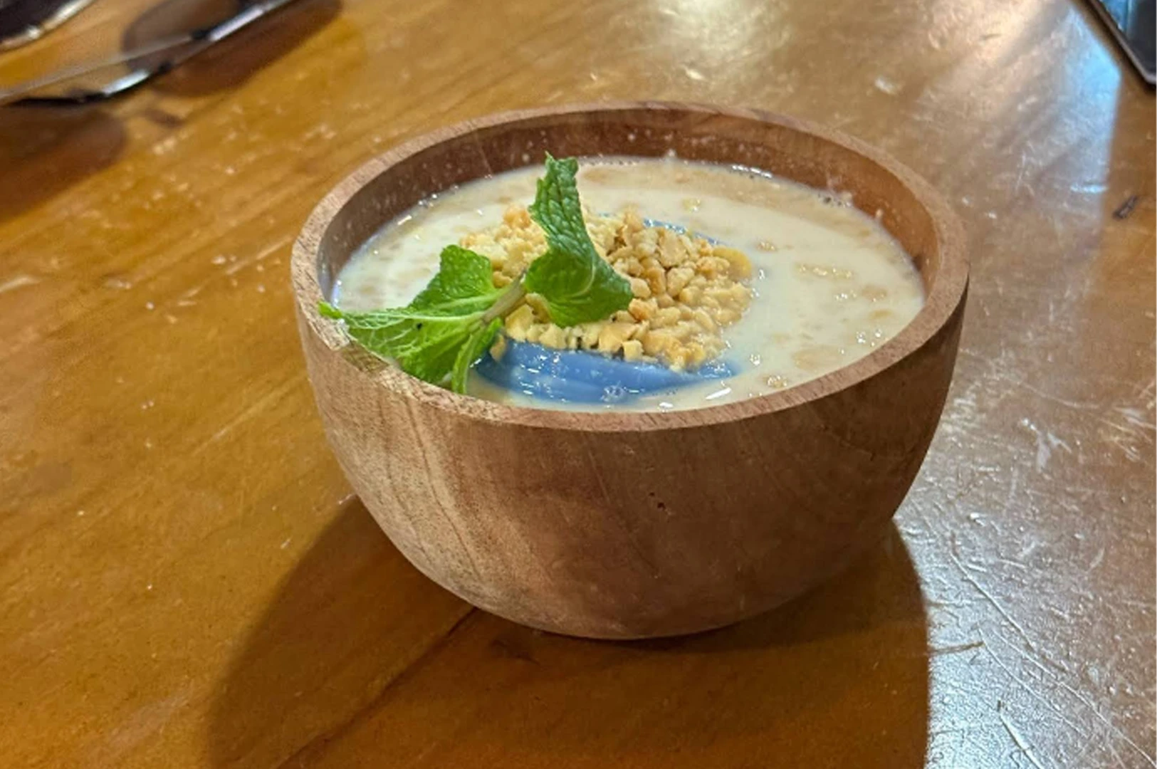 A close-up of a blue pea chia seed pudding served in a rustic wooden bowl on a light-colored wooden table. The creamy white pudding is topped with a swirl of bright blue pea flower extract, a generous sprinkle of crushed peanuts, and a fresh green mint sprig.