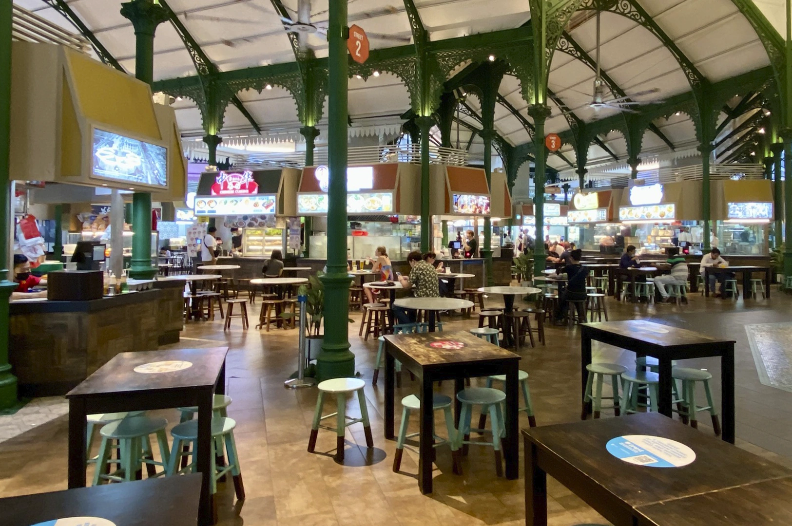 A spacious, high-ceilinged food hall with ornate green iron pillars, featuring various circul