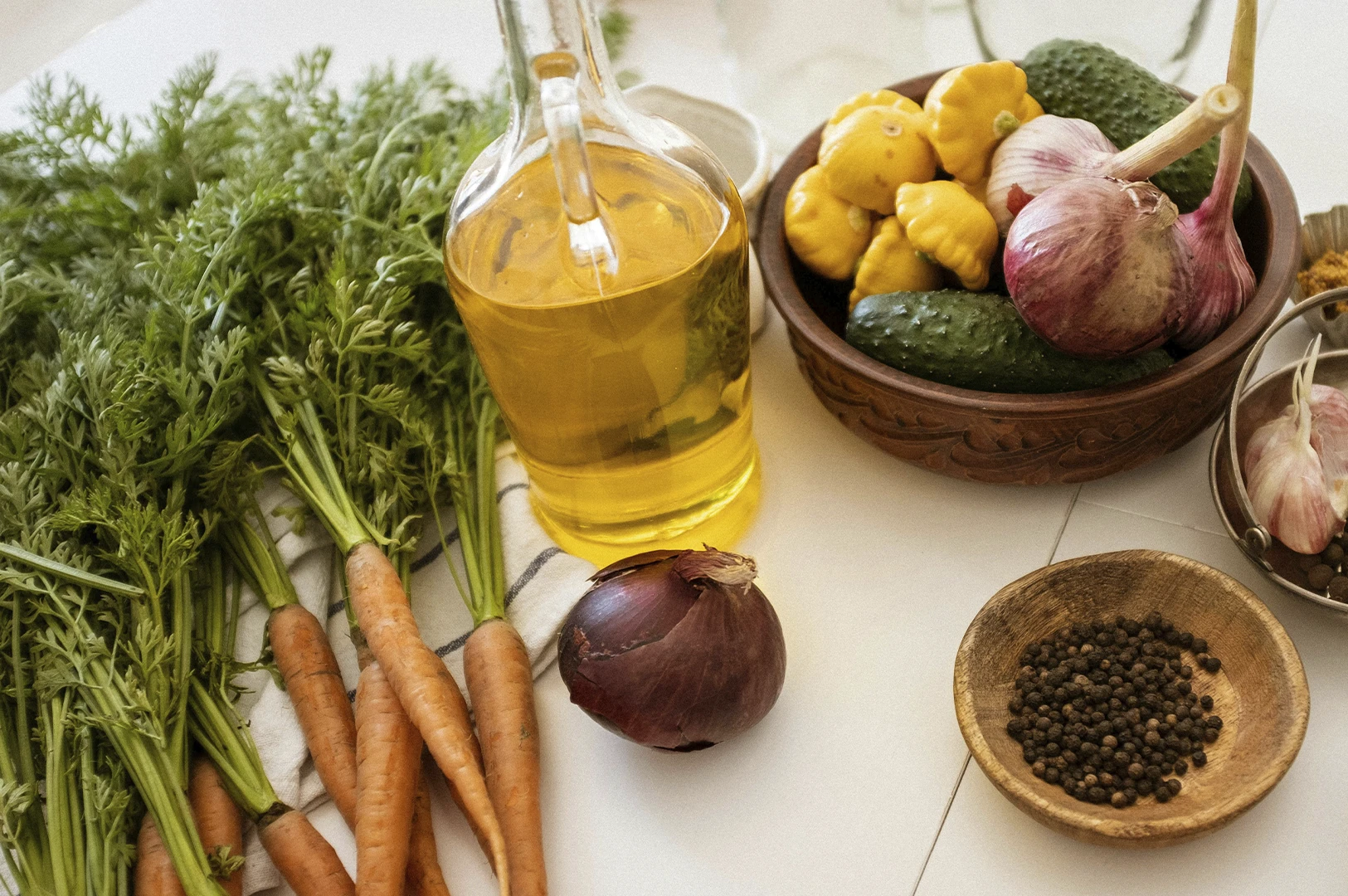 A top-down, brightly lit still life of various fresh cooking ingredients on a white surface. On the left, a large bunch of vibrant green carrot tops sits next to several whole carrots. In the center, a clear glass bottle is filled with golden cooking oil. To the right, a dark clay bowl holds yellow pattypan squash, cucumbers, and a large head of garlic with purple skin. Small wooden and metal bowls containing black peppercorns and ground spices are scattered nearby, resting on a white striped linen cloth.