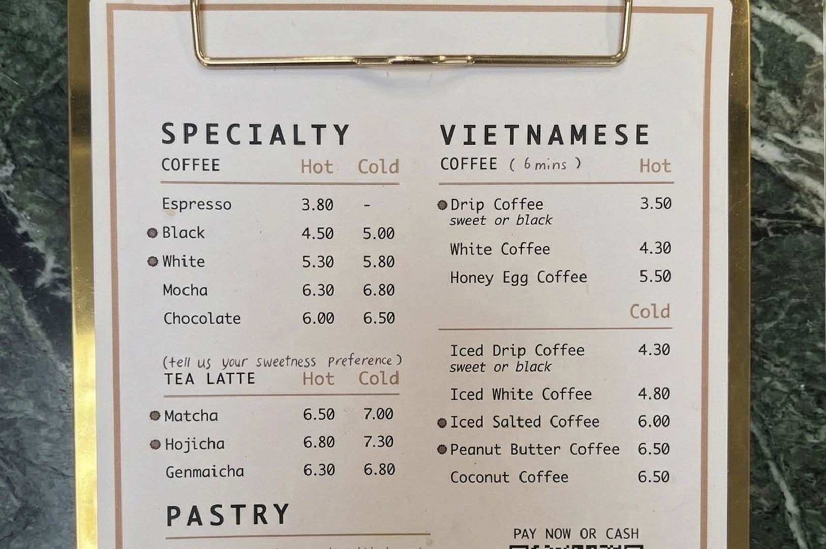 A top-down, clear shot of a printed menu clipped to a gold metal clipboard, resting on a green marble surface. The menu is divided into "Specialty" and "Vietnamese" sections. Under Vietnamese coffee, unique options like "Honey Egg Coffee," "Iced Salted Coffee," and "Peanut Butter Coffee" are listed with their prices. The layout is clean and minimalist, using a sans-serif font with small circular bullet points next to certain items.