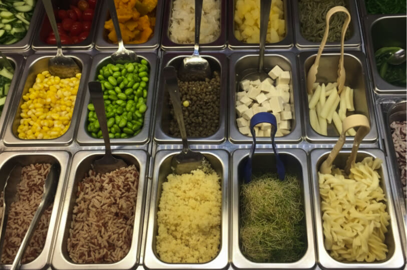 A top-down view of a salad bar featuring stainless steel containers filled with corn, edamame, tofu, pasta, sprouts, and grains.