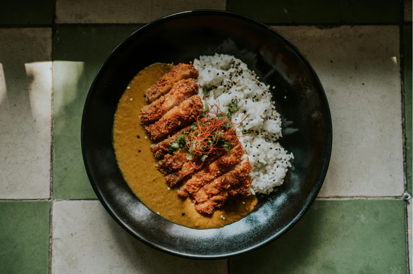 A black bowl of katsu curry and rice viewed from above, garnished with green onions and shredded red chili.