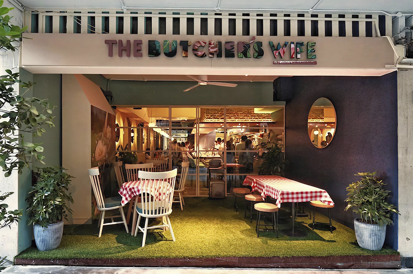 The storefront of a restaurant named "The Butcher's Wife," featuring a cozy outdoor seating area with red-and-white checkered tablecloths and faux grass flooring.
