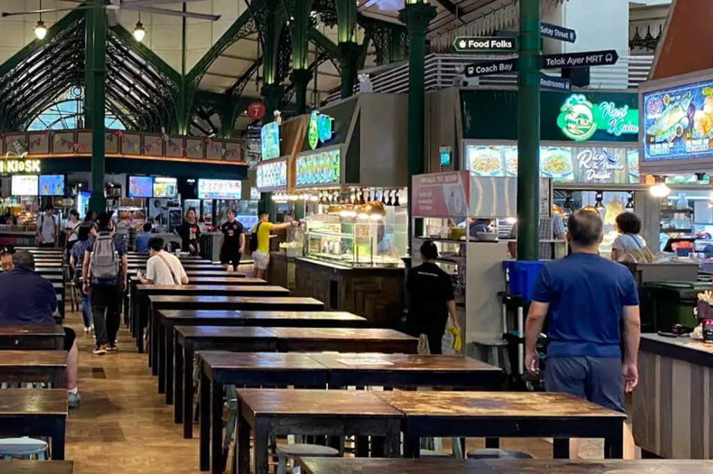 A wide interior shot of Lau Pa Sat hawker centre in Singapore, showing rows of dark wooden communal tables and stools. The architecture features iconic green Victorian-style cast-iron columns and high ceilings. In the background, various food stalls with brightly lit signs, such as "Nasi Katok" and "Rice & Noodle Delight," are visible. People are seen walking through the aisles or seated at tables, capturing the busy, authentic atmosphere of the food hall.