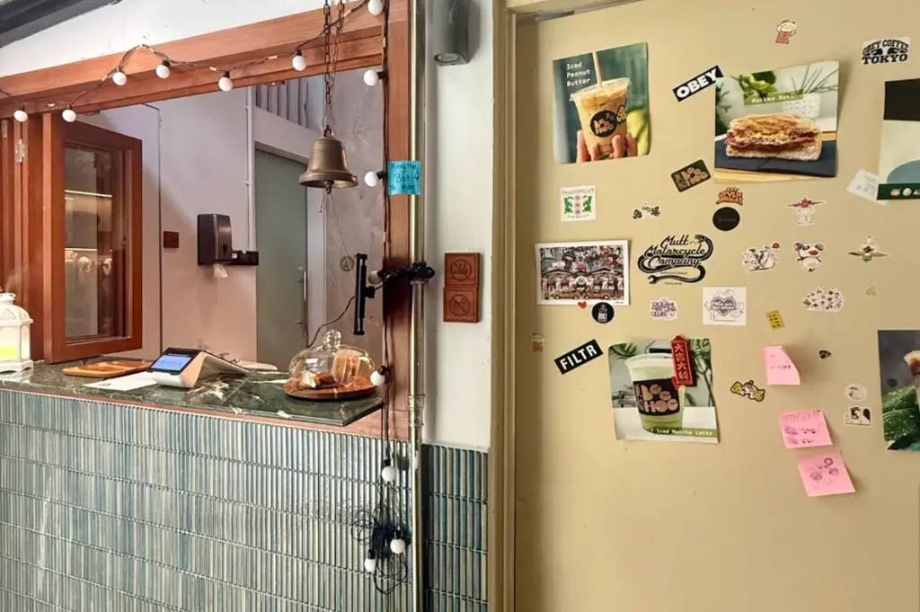 An interior shot of the service counter featuring a teal-colored vertical kit-kat tile backsplash and a dark green marble countertop. To the left, a wooden-framed window is draped with warm string lights and a small brass service bell hangs nearby. To the right, a cream-colored door is covered in various lifestyle stickers, polaroids of food, and pink sticky notes. A small glass cloche on the counter holds a pastry, contributing to the cozy, eclectic aesthetic of the cafe.