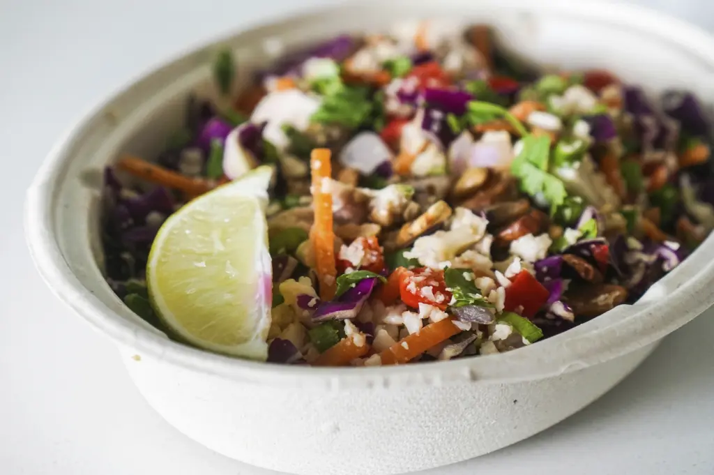 A close-up, slightly angled shot of a colorful grain bowl served in a white compostable paper bowl. The dish is packed with shredded red cabbage, chopped carrots, peanuts, cilantro, and white grains, topped with a fresh lime wedge on the side. The focus is sharp on the textures of the vegetables in the foreground, while the white background is clean and minimalist.