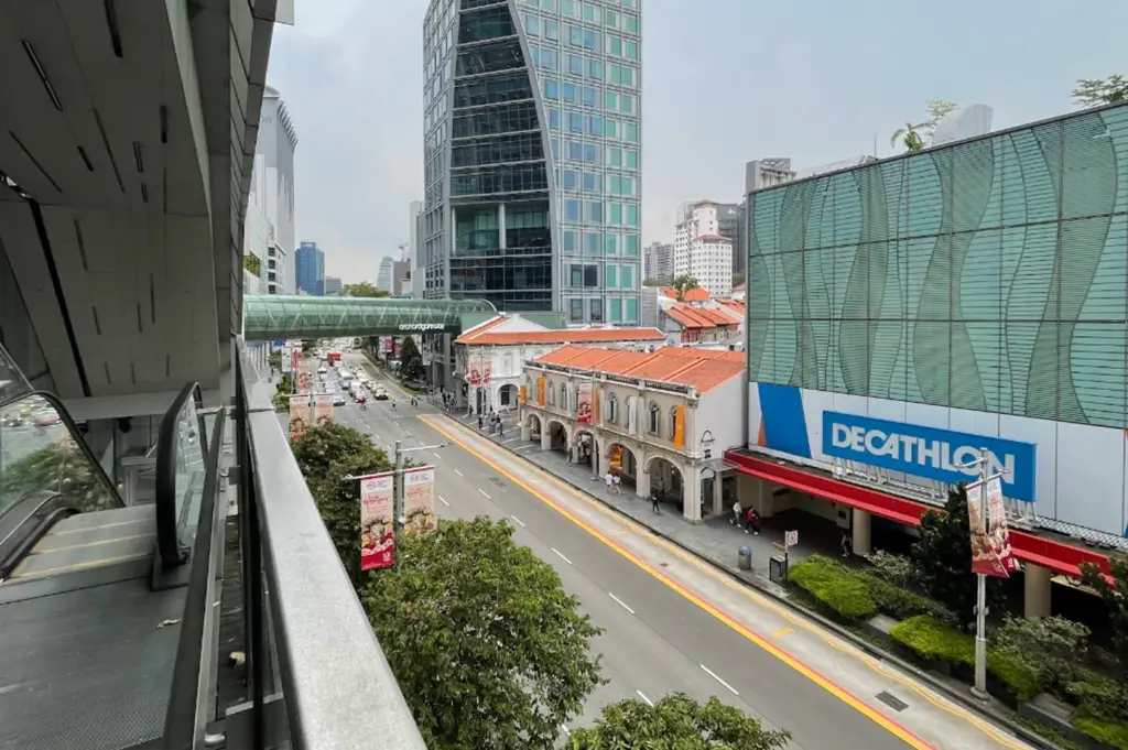 An elevated view looking down Orchard Road. On the right, a large Decathlon store is visible with its signature blue and white branding. Next to it are traditional two-story shophouses with terracotta tiled roofs and arched walkways. In the background, a modern glass skyscraper with a unique geometric "cut-out" design towers over the street, and a green covered pedestrian bridge spans across the road.