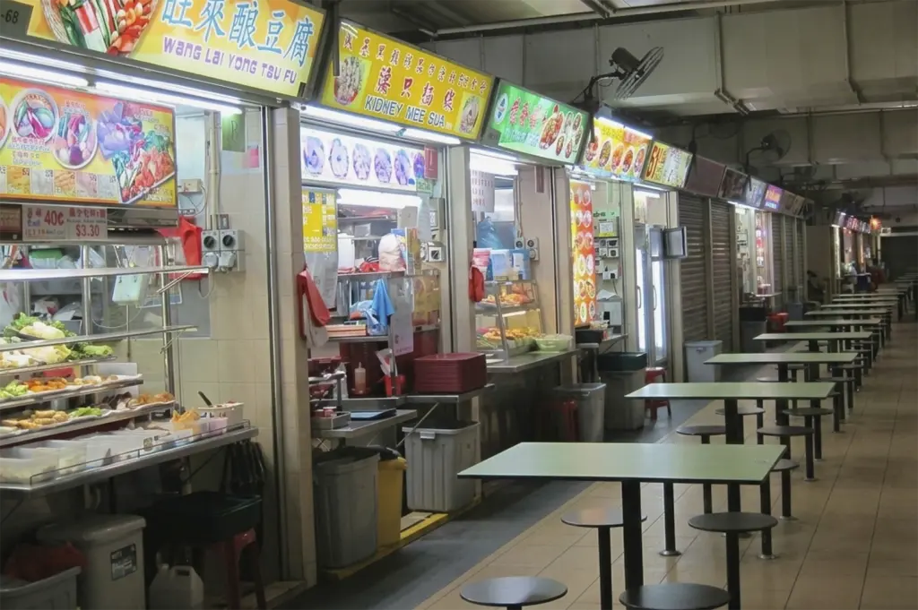 An eye-level view of a quiet hawker center aisle lined with food stalls featuring bright yellow and green signage, including stalls for "Yong Tau Fu" and "Kidney Mee Sua," next to rows of empty green tables and stools.