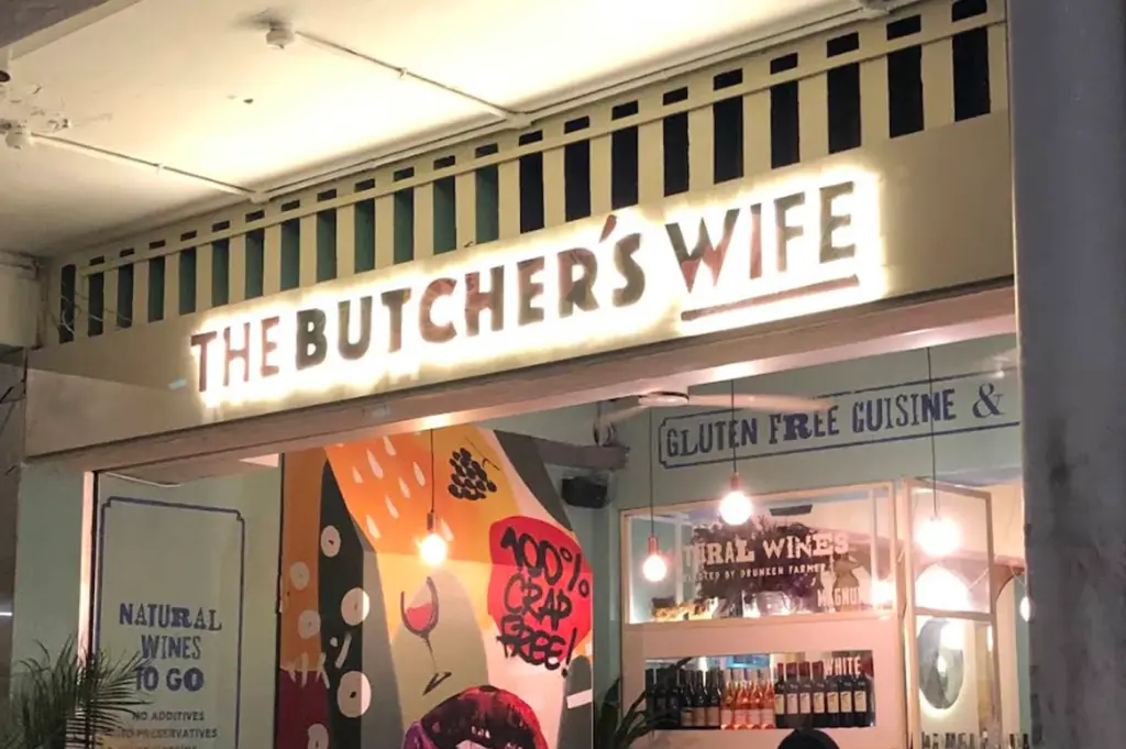 A night-time, eye-level shot of the storefront for a restaurant named "The Butcher's Wife." The main signage features bold, backlit white lettering against a pale yellow facade with a black-and-white striped decorative border above. Below the sign, the interior is visible through a large open entrance, showing a vibrant mural with abstract shapes, a wine glass illustration, and the text "100% Crap Free!" in a red circle. To the right, a window display features bottles of wine and a sign for "Natural Wines." A blue-bordered sign on the left reads "Natural Wines To Go," and another sign above the window mentions "Gluten Free Cuisine." Warm, exposed light bulbs hang from the ceiling, creating a welcoming glow.