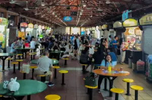 A wide-angle, eye-level shot capturing the bustling interior of Maxwell Food Centre. Rows of green and orange circular tables with matching yellow stools fill the foreground and middle ground, occupied by diverse groups of diners. Above, the high, industrial timber-truss ceiling is lined with white ceiling fans and rows of fluorescent lights. Two long aisles are flanked by brightly lit hawker stalls with colorful signage. A man in a light blue shirt sits at an orange table in the lower right, looking toward the camera.