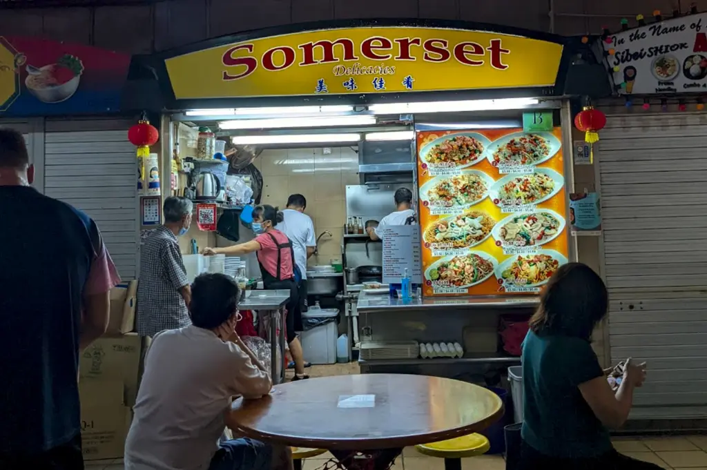 A brightly lit hawker stall named Somerset Delicacies (美味佳肴) under a yellow curved sign. The stall is located in a row of shops with metal shutters. A large, colorful menu board on the right displays photos of various fried rice and noodle dishes. Two staff members are visible inside the small kitchen area, and customers are seated at a round wooden table in the foreground.