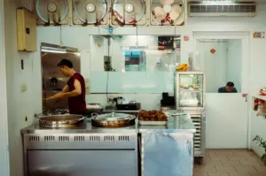 Asian street food stall kitchen interior with cook preparing dishes at stainless steel counter, overhead ventilation fans, glass display of roasted meats and clean tiled workspace in a eatery setting.