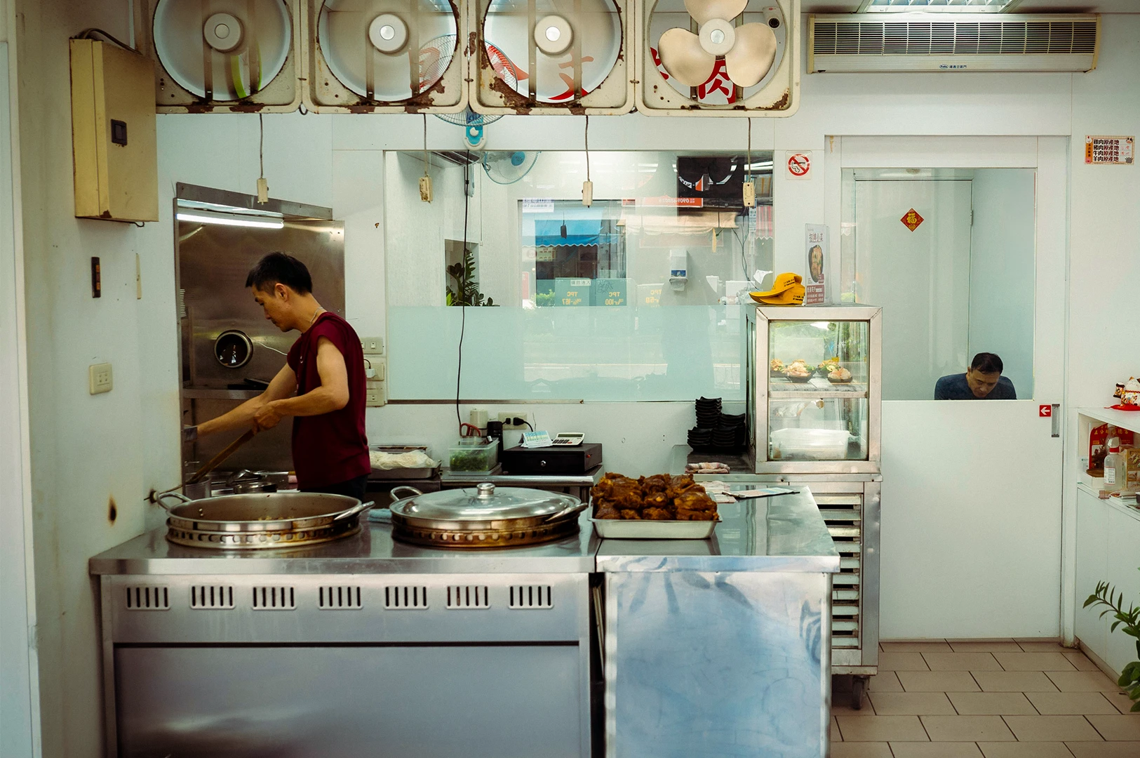 Asian street food stall kitchen interior with cook preparing dishes at stainless steel counter, overhead ventilation fans, glass display of roasted meats and clean tiled workspace in a eatery setting.