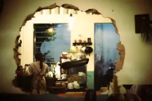 A barista in a striped shirt and apron working behind a coffee bar, viewed through a large, jagged hole in a white interior wall.
